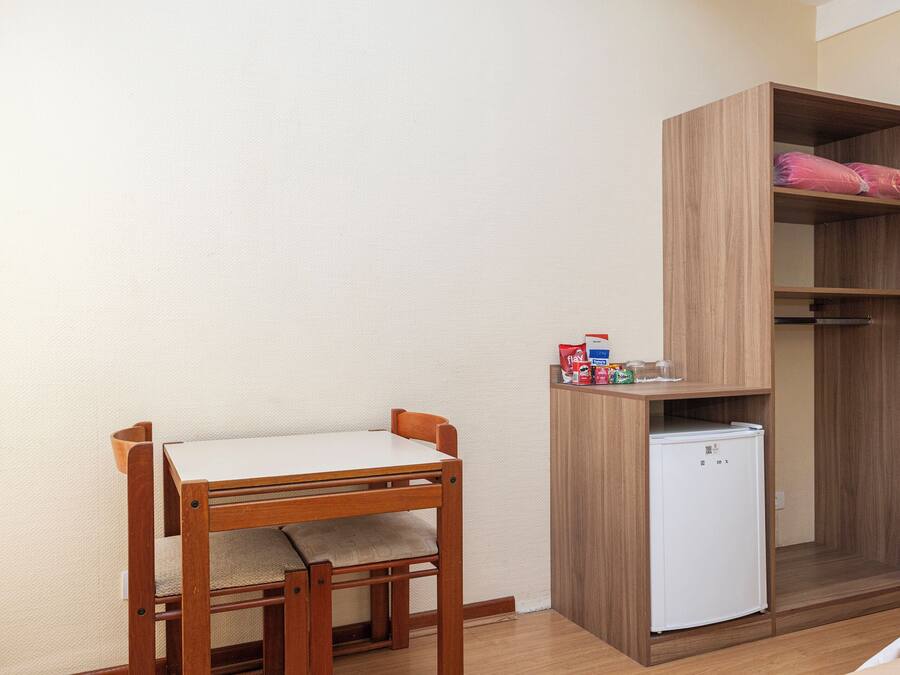 Una habitación compacta con una mesa y sillas de madera, un refrigerador blanco pequeño y un armario de madera con un cojín rosa.