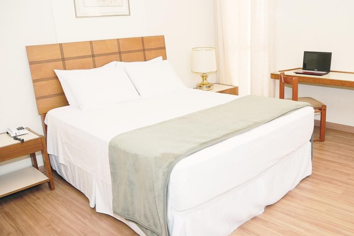 A neatly made bed with a headboard, a bedside table with a lamp, and a desk with a laptop.