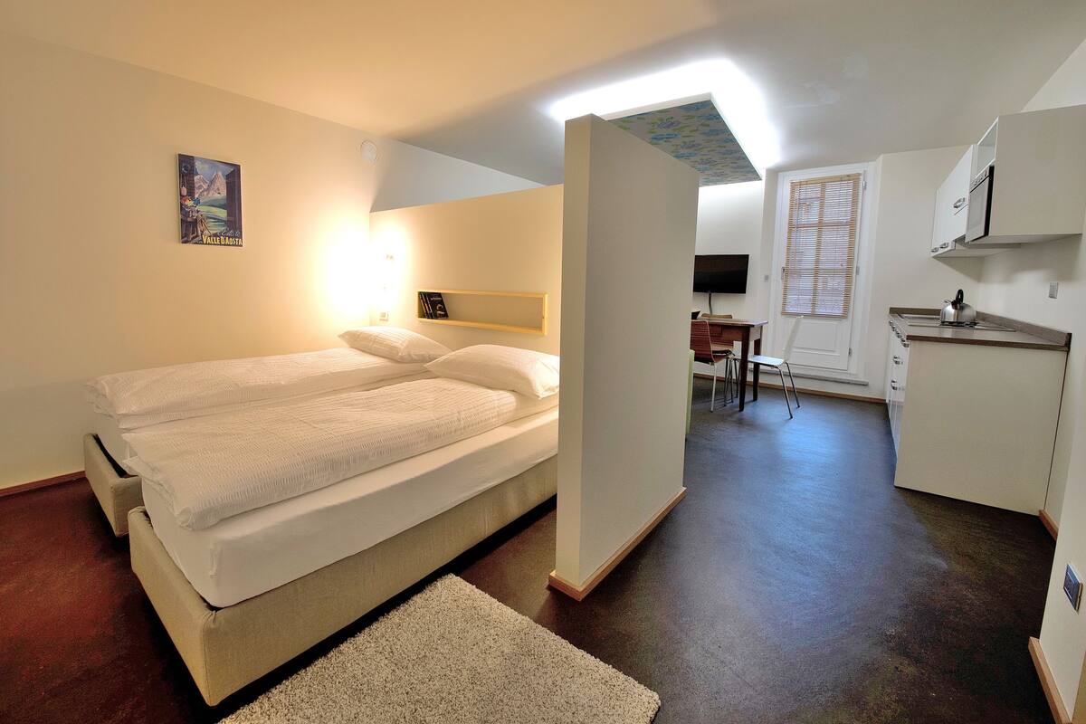 A bedroom with a bed, a desk, and a kitchenette.