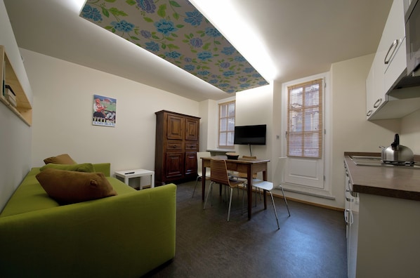 A compact living space with a green sofa, wooden cabinet, dining table, and a kitchenette.