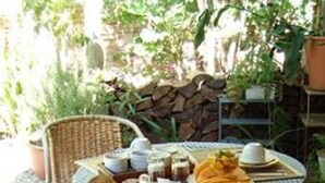 An outdoor breakfast setup with a wicker chair, a table with checkered tablecloths, plates of food, and a jug of orange juice. - Colonia Suite Apartments (Colonia del Sacramento)