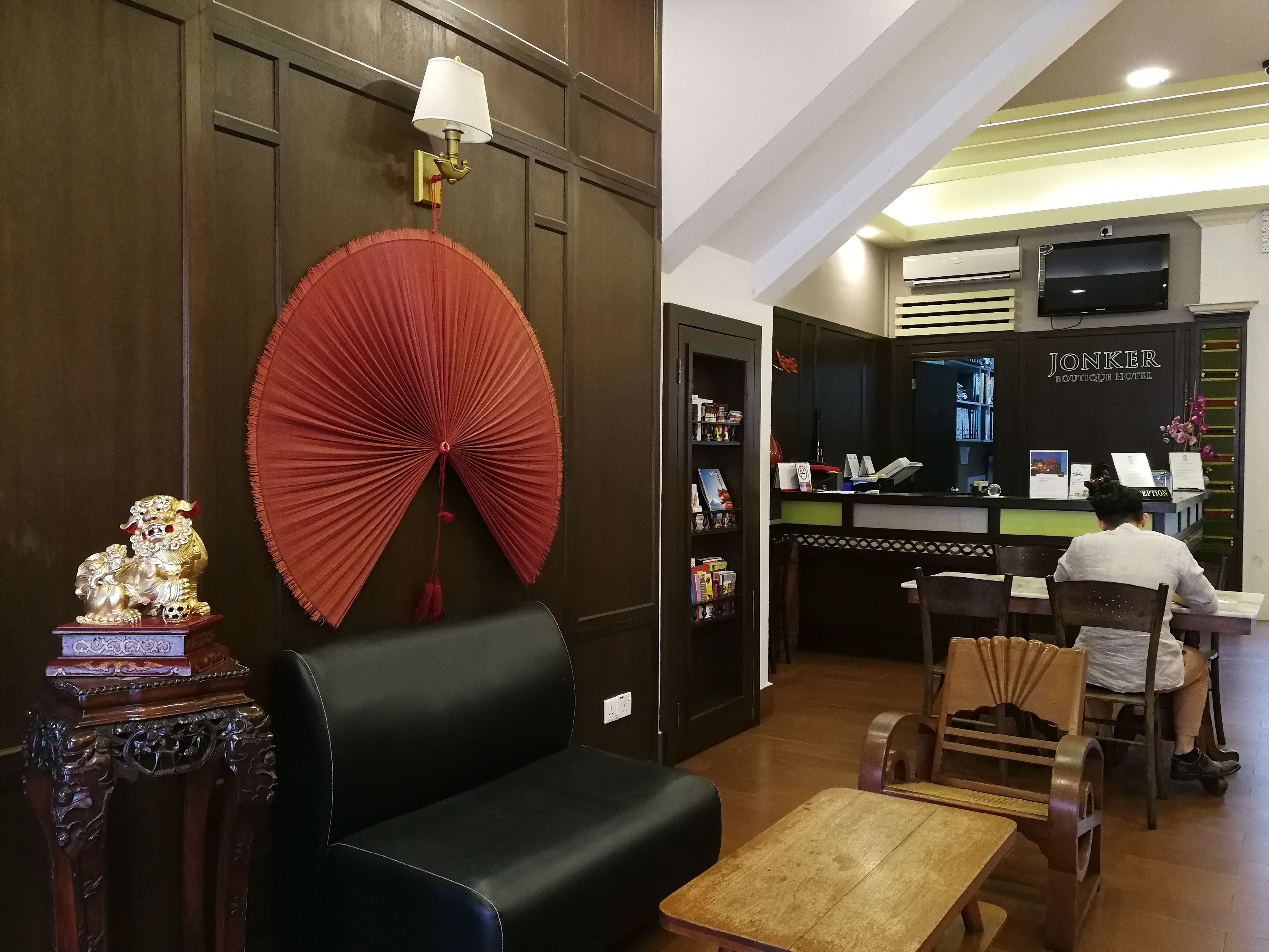 Zona de recepción de un hotel con un mostrador, un sofá de cuero negro, un banco de madera y un abanico decorativo rojo.
