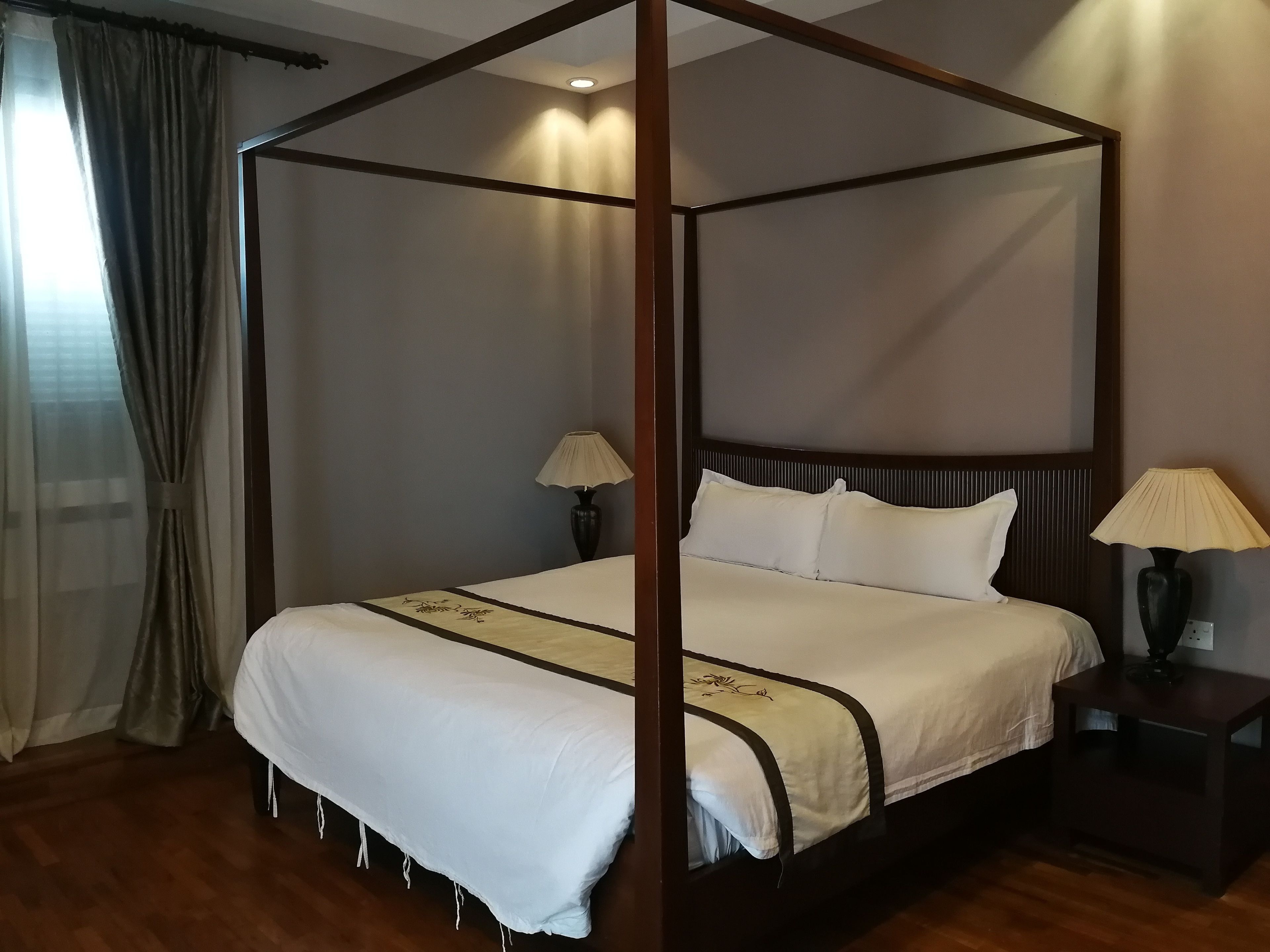 Una cama con dosel tapizada con sábanas blancas y un borde decorativo, flanqueada por dos mesitas de noche con lámparas, en una habitación con suelo de madera y una paleta de colores neutros.