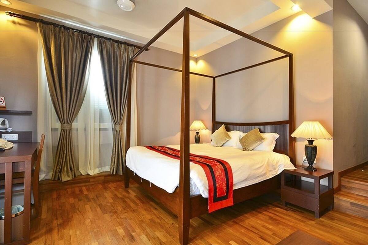 A four-poster bed with a wooden frame, white bedding, and a red throw blanket. There are two bedside tables with lamps, a wooden desk, and a chair. The room has wooden flooring and a large window with curtains.