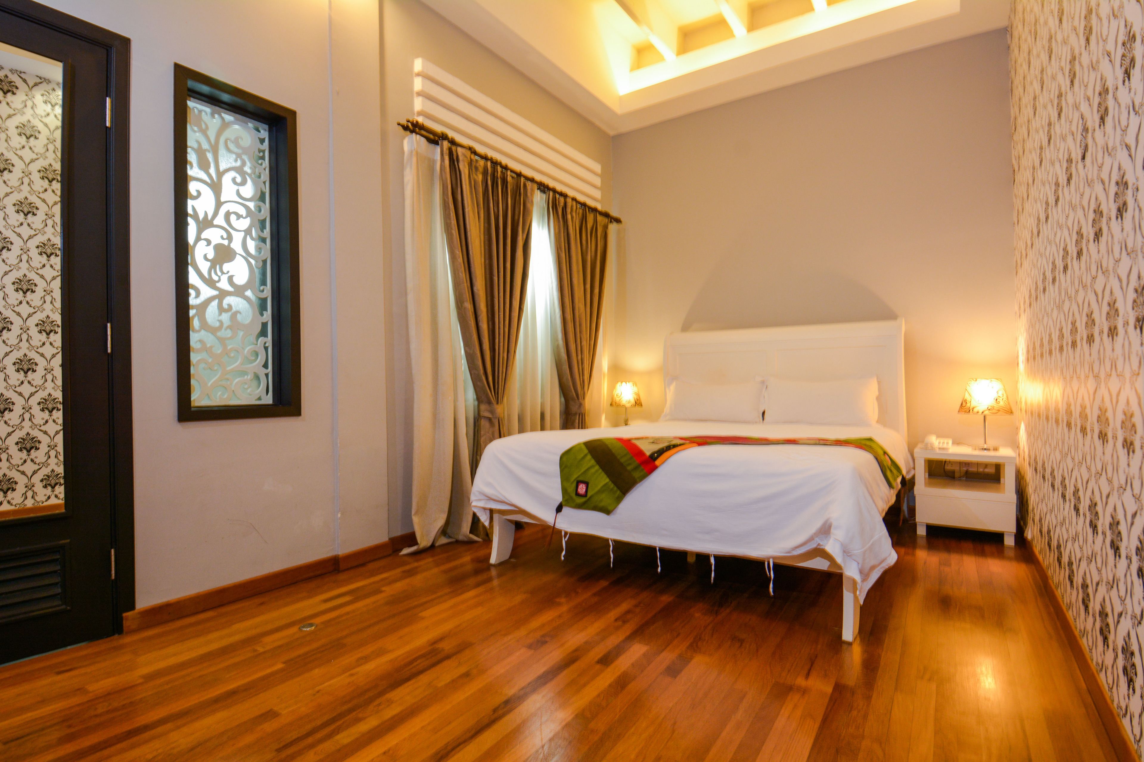 A hotel room with a wooden floor, a bed with white bedding, a nightstand with a lamp, and a decorative wall panel.