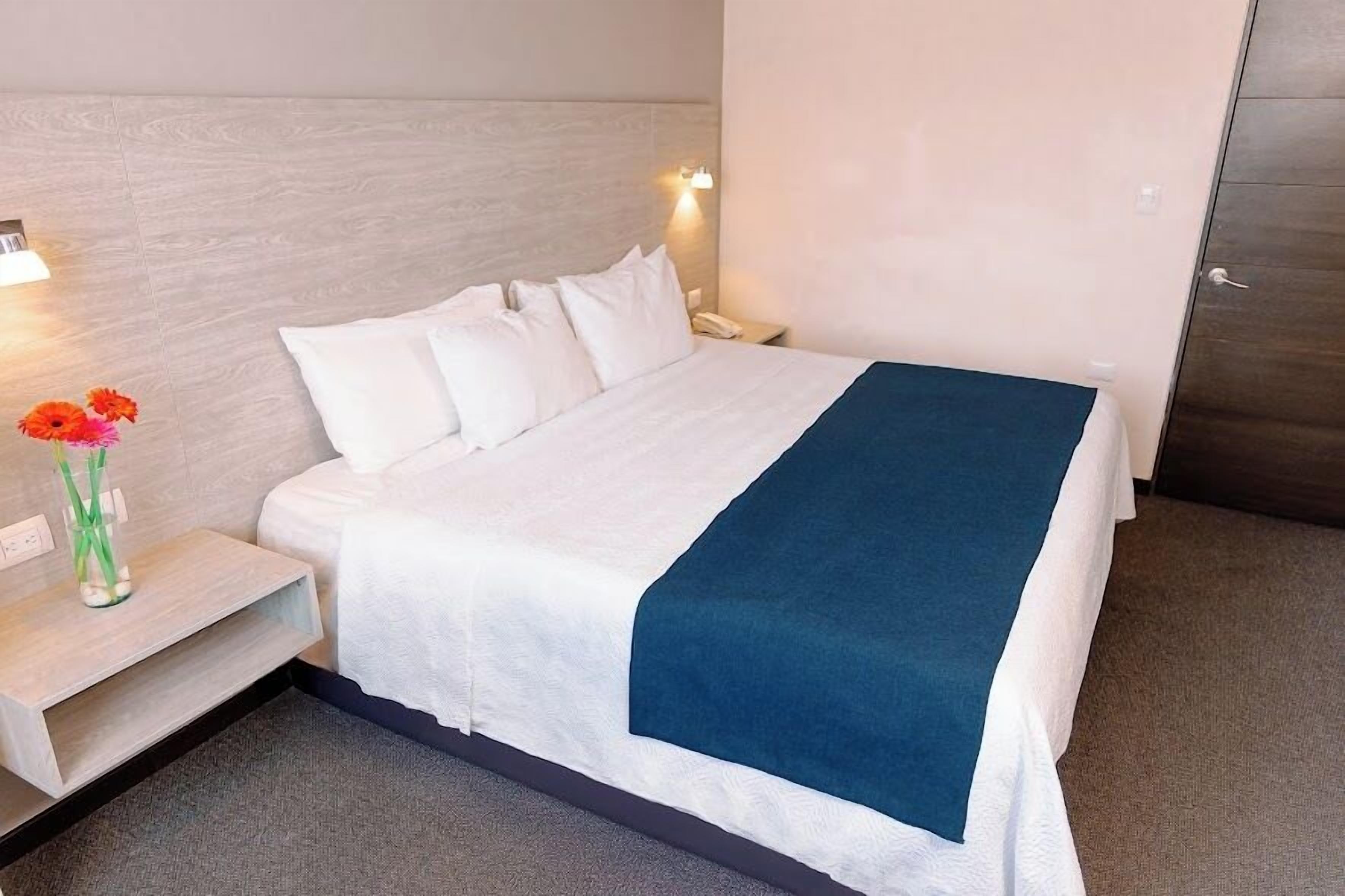 Habitación de hotel con cama, mesita de noche, un jarrón con flores y una lámpara de pared.
