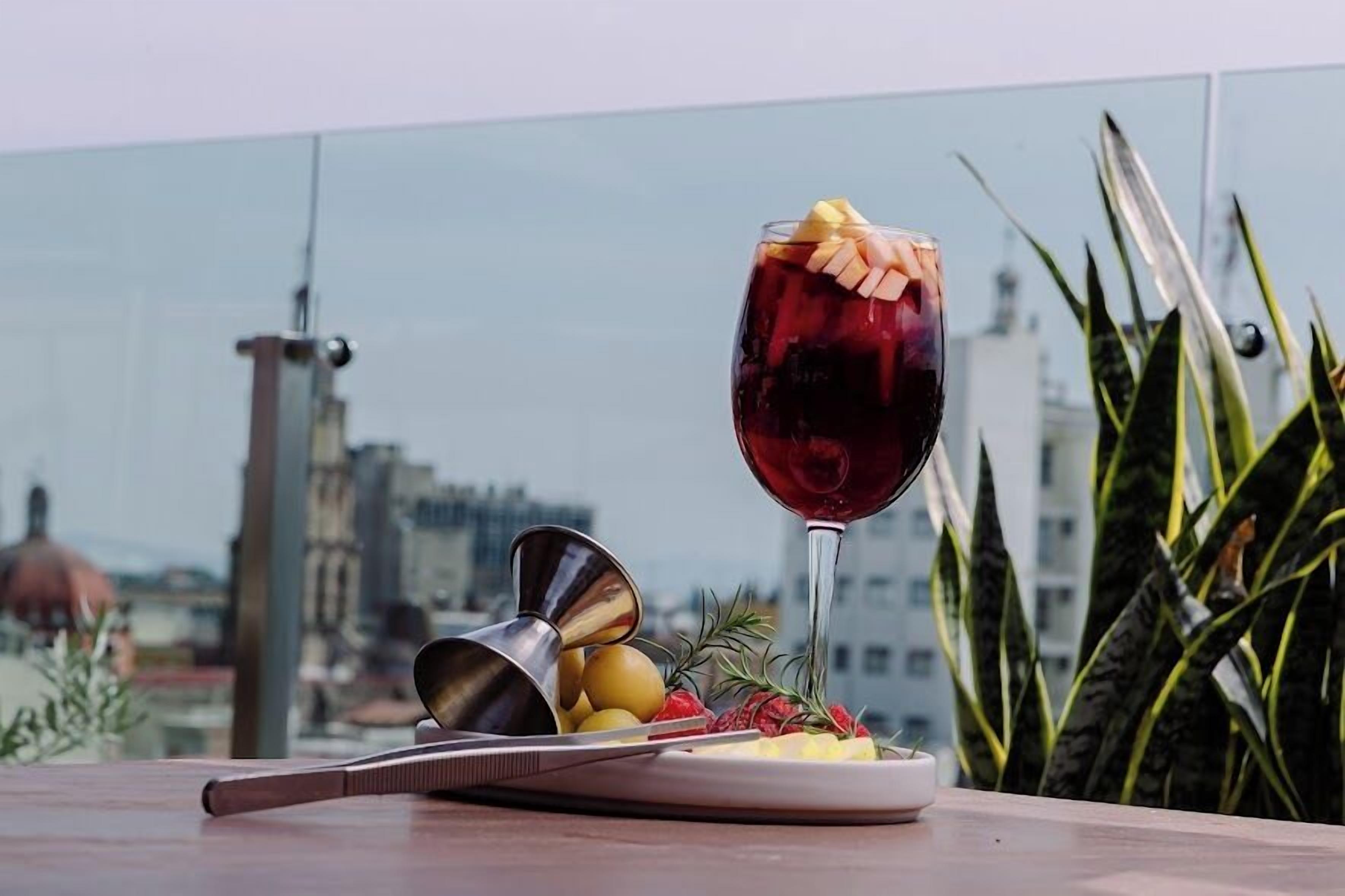 A glass of red wine on a table with a cityscape view, a plate of fruit, and a metal pitcher.