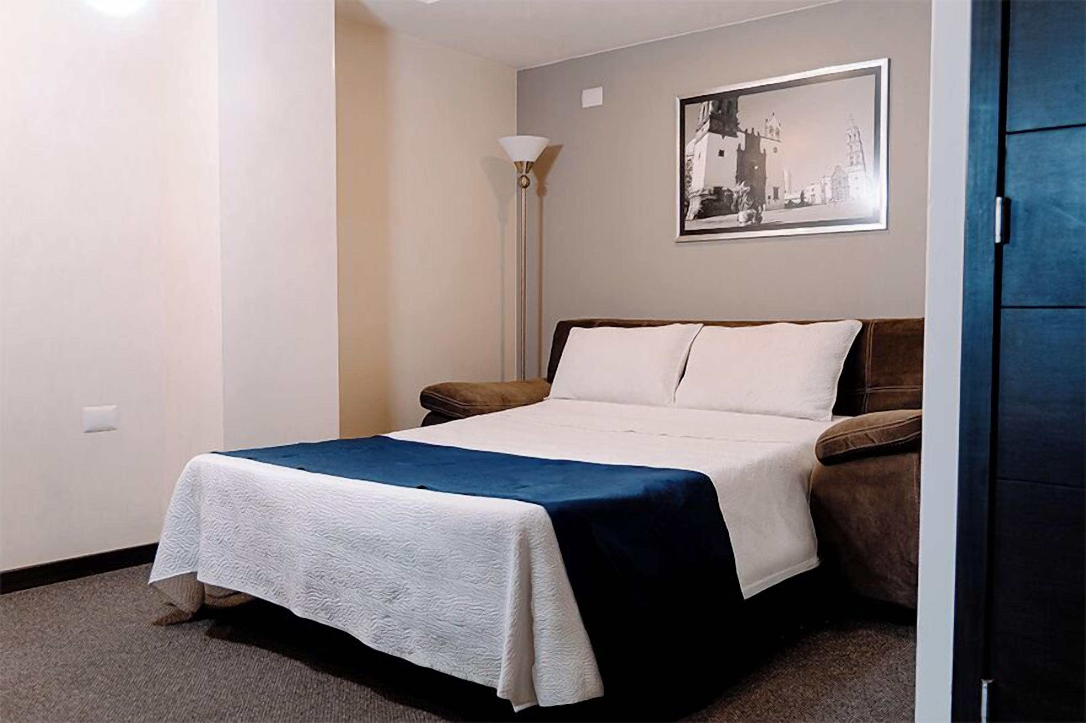 Un cuarto de hotel con una cama, un sillón marrón, una lámpara de pie y un cuadro en la pared.