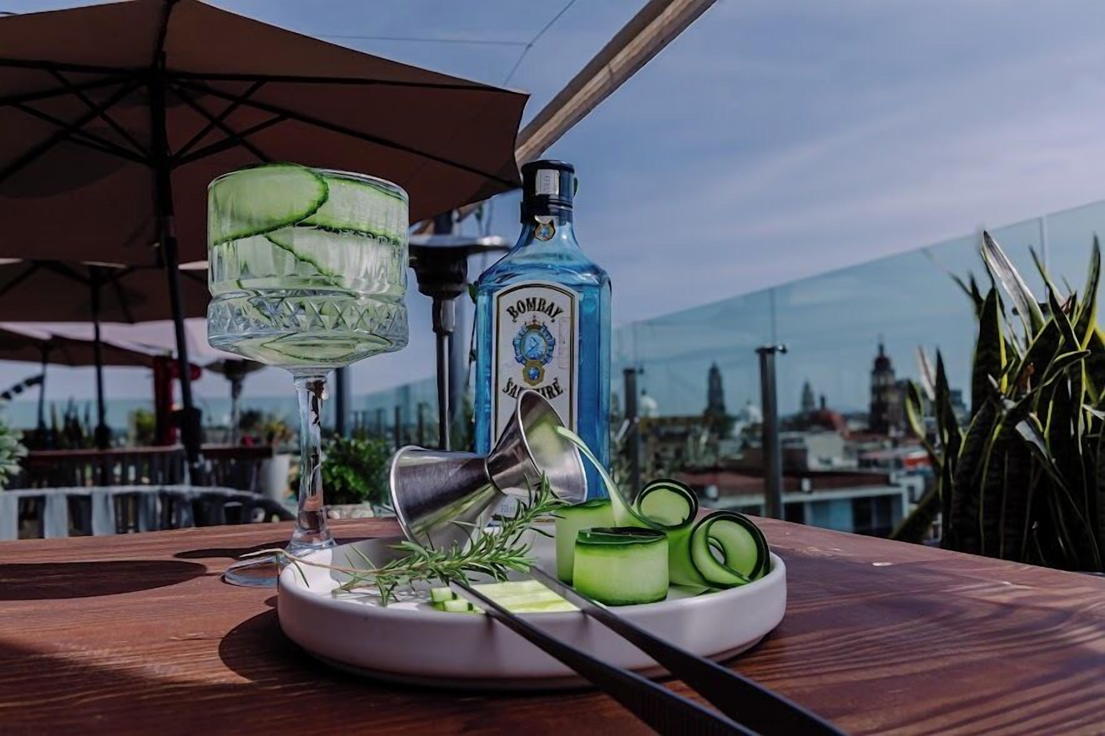 Un cocktail agrémenté de rondelles de concombre, une bouteille de gin Bombay Sapphire et un shaker à cocktail, le tout posé sur une table avec une vue sur la ville en arrière-plan.