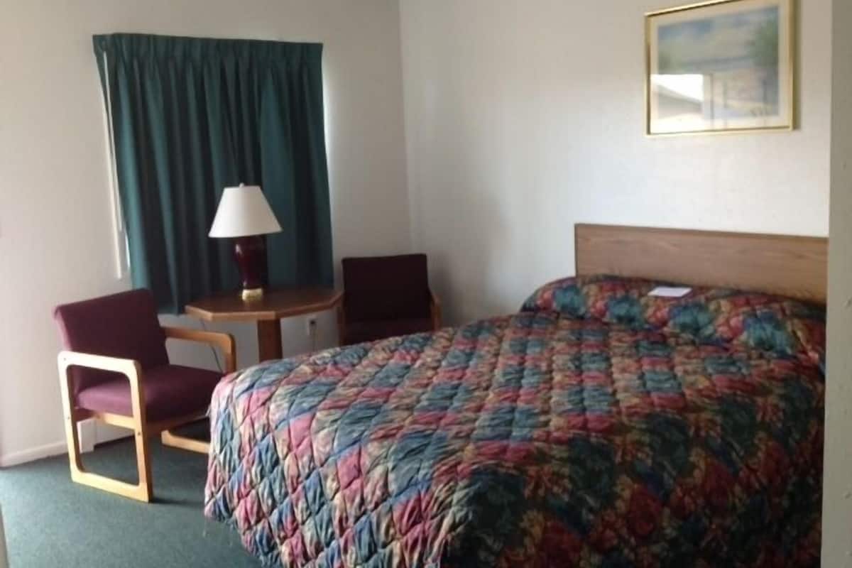 A hotel room with a bed, a desk, two chairs, a lamp, and a painting on the wall.