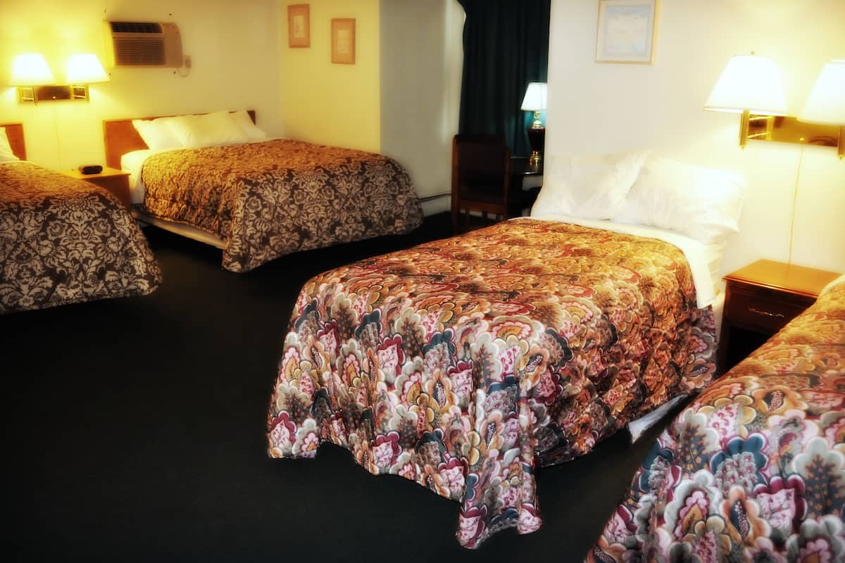 A hotel room with three beds, each with a patterned bedspread, a bedside table with a lamp, and a wall-mounted air conditioning unit.