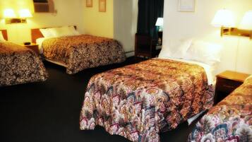 A hotel room with three beds, each with a patterned bedspread, a bedside table with a lamp, and a wall-mounted air conditioning unit.