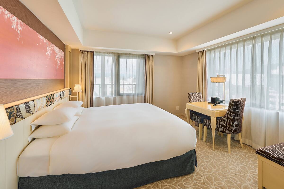 A hotel room with a large bed, a desk with a chair, and a view of the city through the windows.