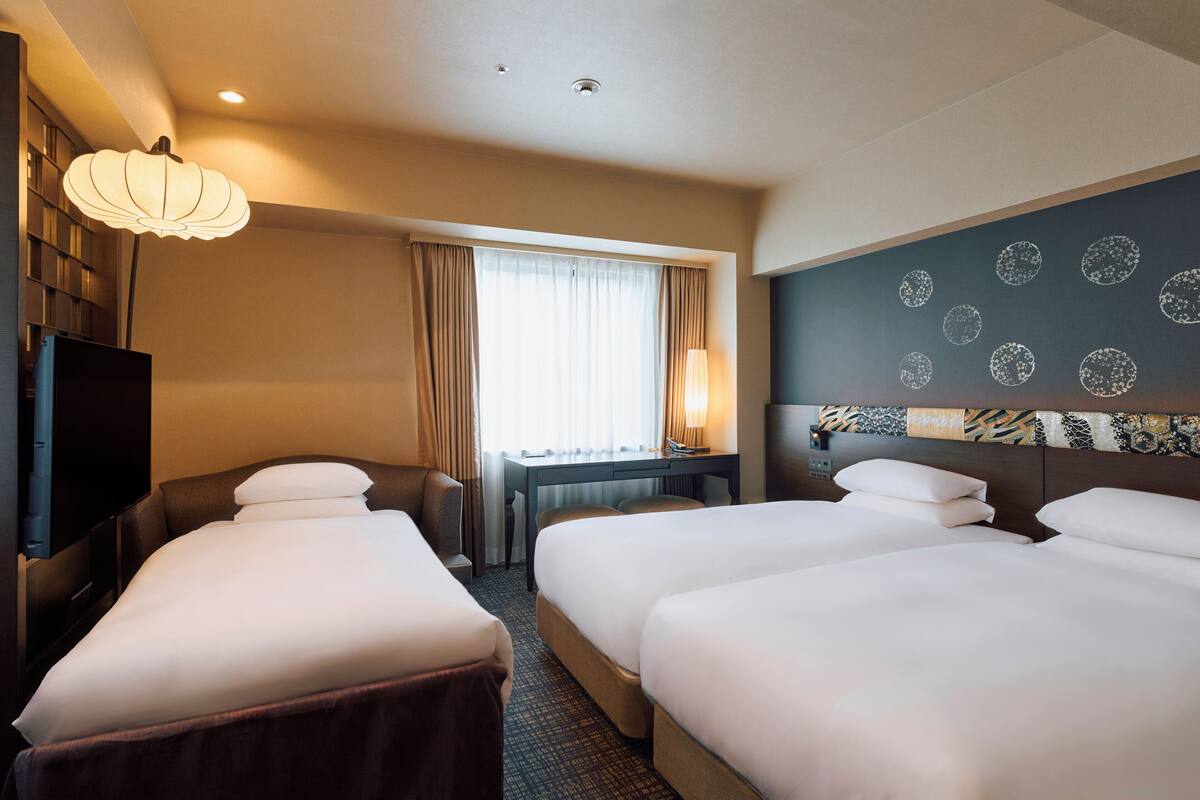 A hotel room with two beds, a desk, a chair, a TV, and a large window with curtains.