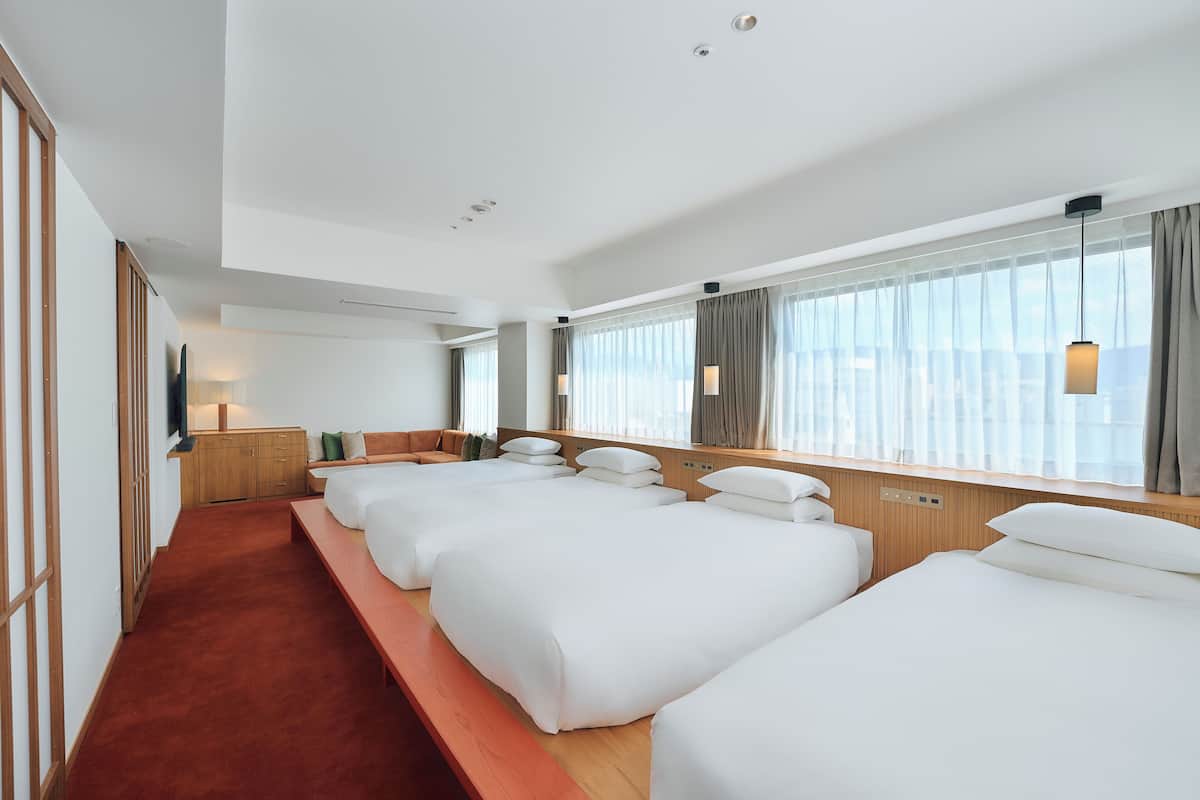 A hotel room with multiple beds, a wooden bench, a TV, and large windows with curtains.