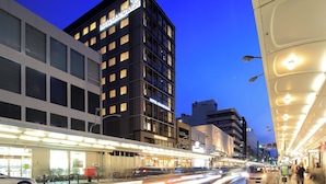 A modern hotel with a prominent 'HOTEL GRAND BACH' sign, a covered pedestrian walkway, and a busy street with vehicles. - Hotel Grand Bach Kyoto Select (Kyoto)