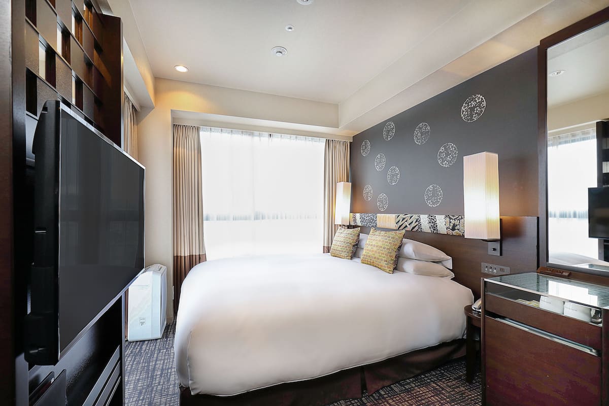 A hotel room with a large bed, a flat-screen TV, a desk with a lamp, and a large window with curtains.