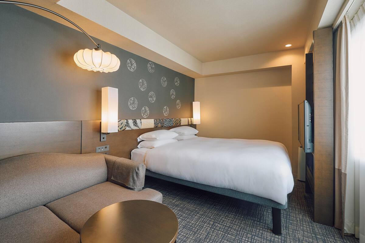 A hotel room with a bed, a sofa, a round table, and a wall-mounted TV.