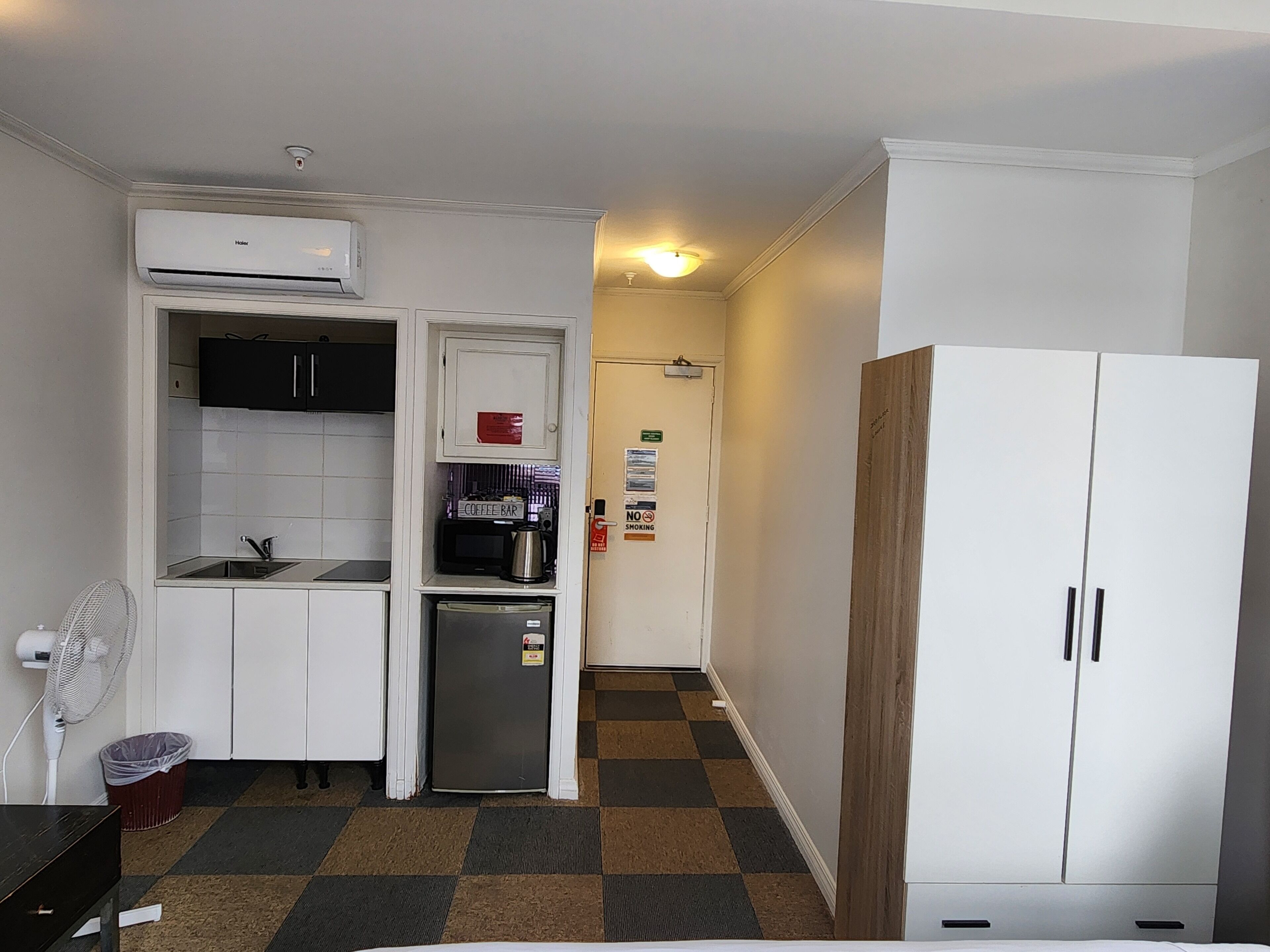 A compact kitchen with a built-in microwave, a coffee maker, and a small refrigerator. There's a white cabinet with a 'No Smoking' sign and a door with a 'Coffee Bar' sign. A fan is on the floor, and a wooden wardrobe is visible.