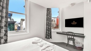 A hotel room with a bed, a TV, a window with a view of buildings, a chair, and a trash bin. - Albion of Auckland (Auckland)