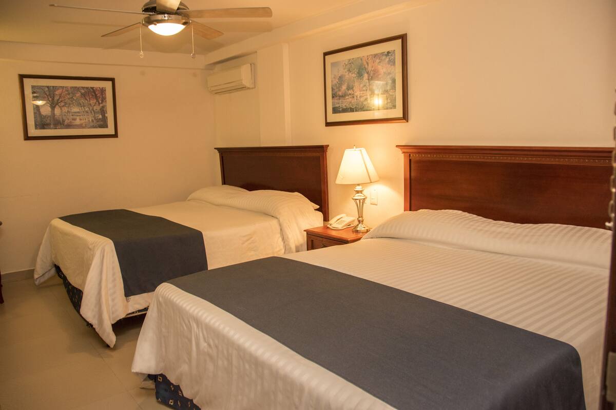 Une chambre d’hôtel avec deux lits simples, un ventilateur de plafond, une lampe de chevet et deux tableaux encadrés au mur.