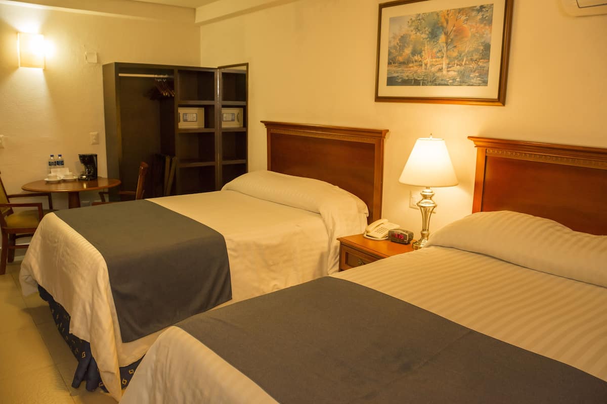 Une chambre d’hôtel avec deux lits, une table de chevet avec une lampe, un bureau avec une chaise, une bibliothèque et un tableau au mur.