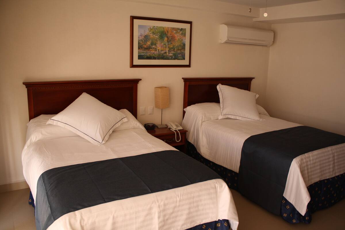Une chambre d’hôtel avec deux lits, chacun doté d’une tête de lit en bois, de draps blancs et d’une couvre-lit bleu foncé. Un tableau encadré est accroché au mur, et un appareil de climatisation est installé au-dessus des lits.