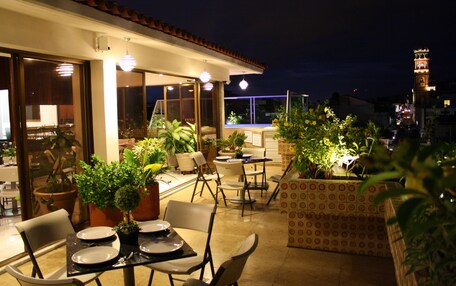 An outdoor dining area with tables and chairs, potted plants, and a view of the cityscape at night.