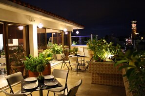 An outdoor dining area with tables and chairs, potted plants, and a view of the cityscape at night.