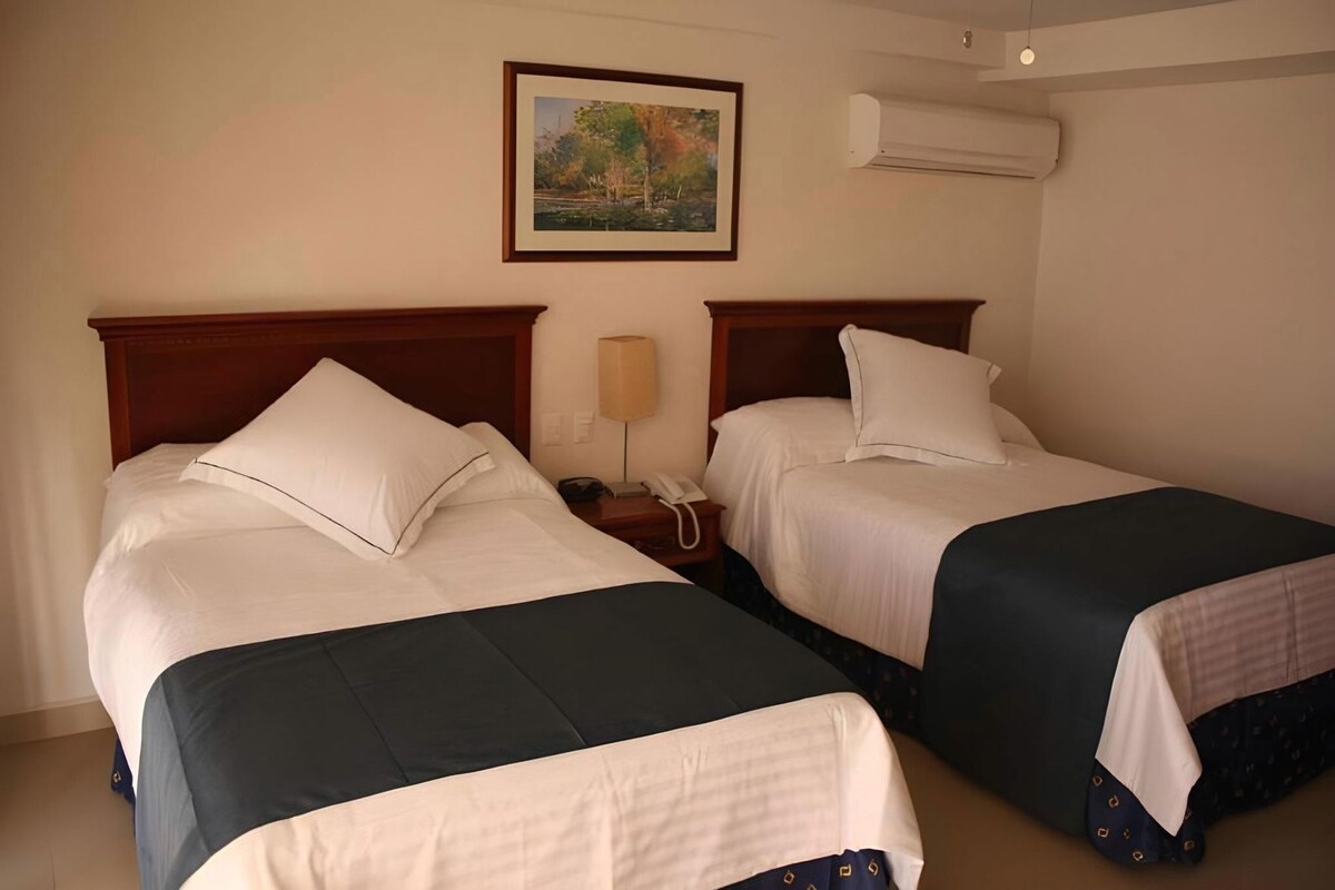 Una habitación de hotel con dos camas, una mesita de noche con lámpara, un teléfono y un cuadro en la pared.