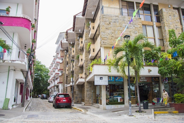 A street with multi-story buildings, a red car parked on the side, and a woman sitting on a bench.