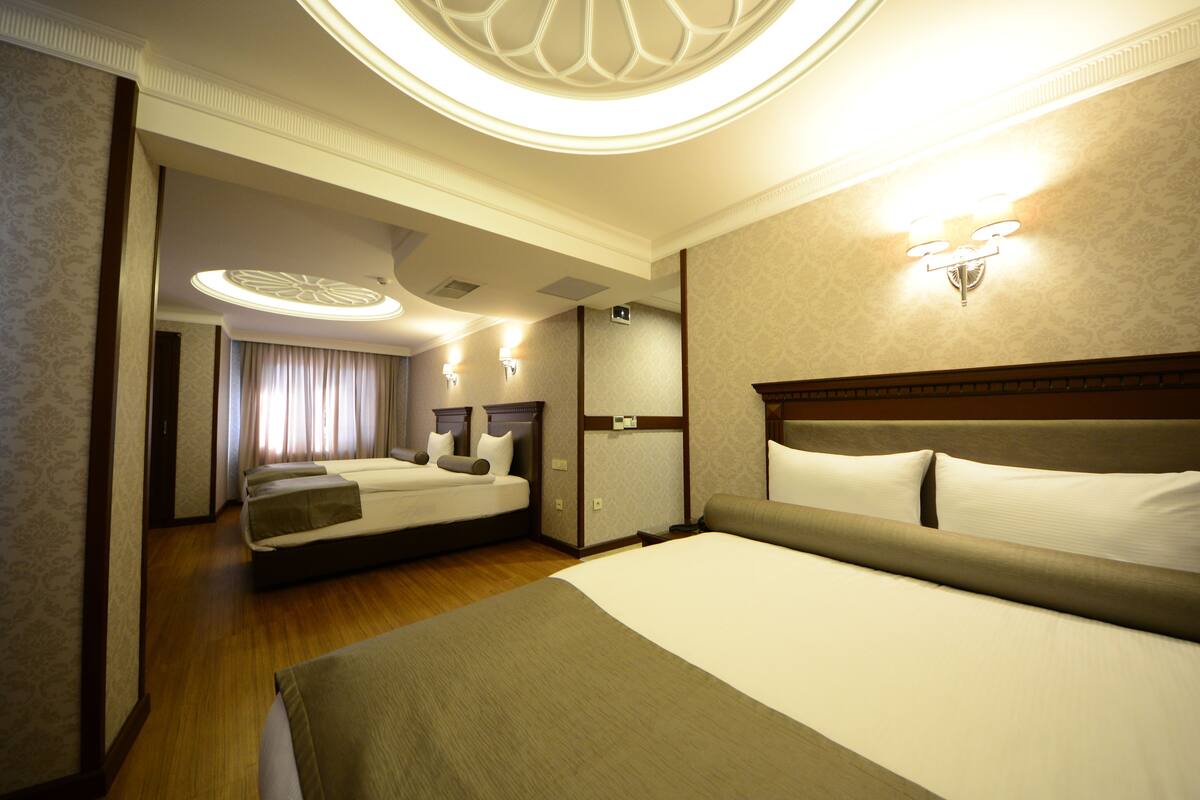 A hotel room with two beds, a large window with curtains, a chandelier, and a wall-mounted light fixture.