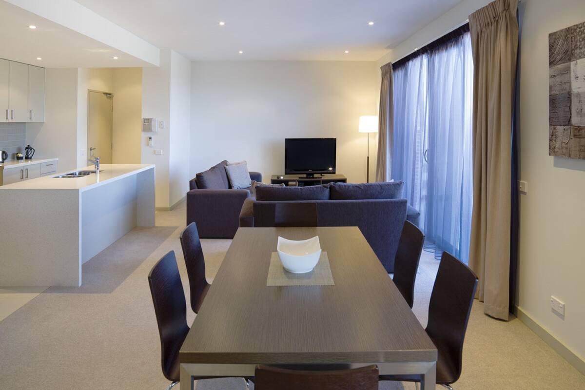 A modern hotel room with a dining area, a sofa, a TV, and a kitchen.
