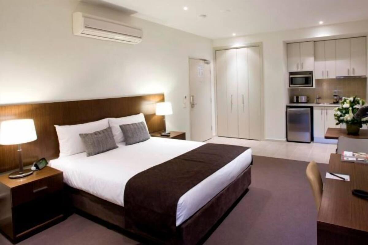 A hotel room with a bed, bedside tables, a desk, a chair, a microwave, a refrigerator, and a door.
