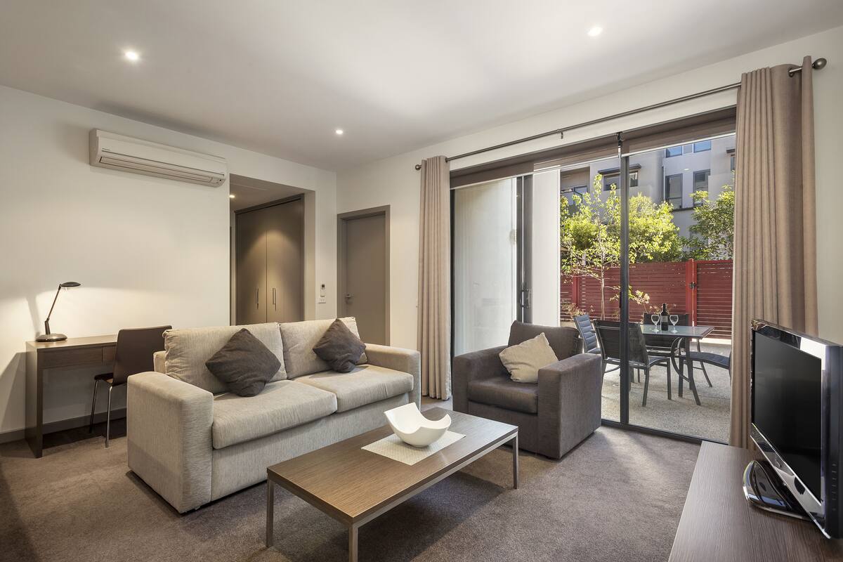 A modern living room with a sofa, armchair, coffee table, TV, and a sliding glass door leading to an outdoor patio.