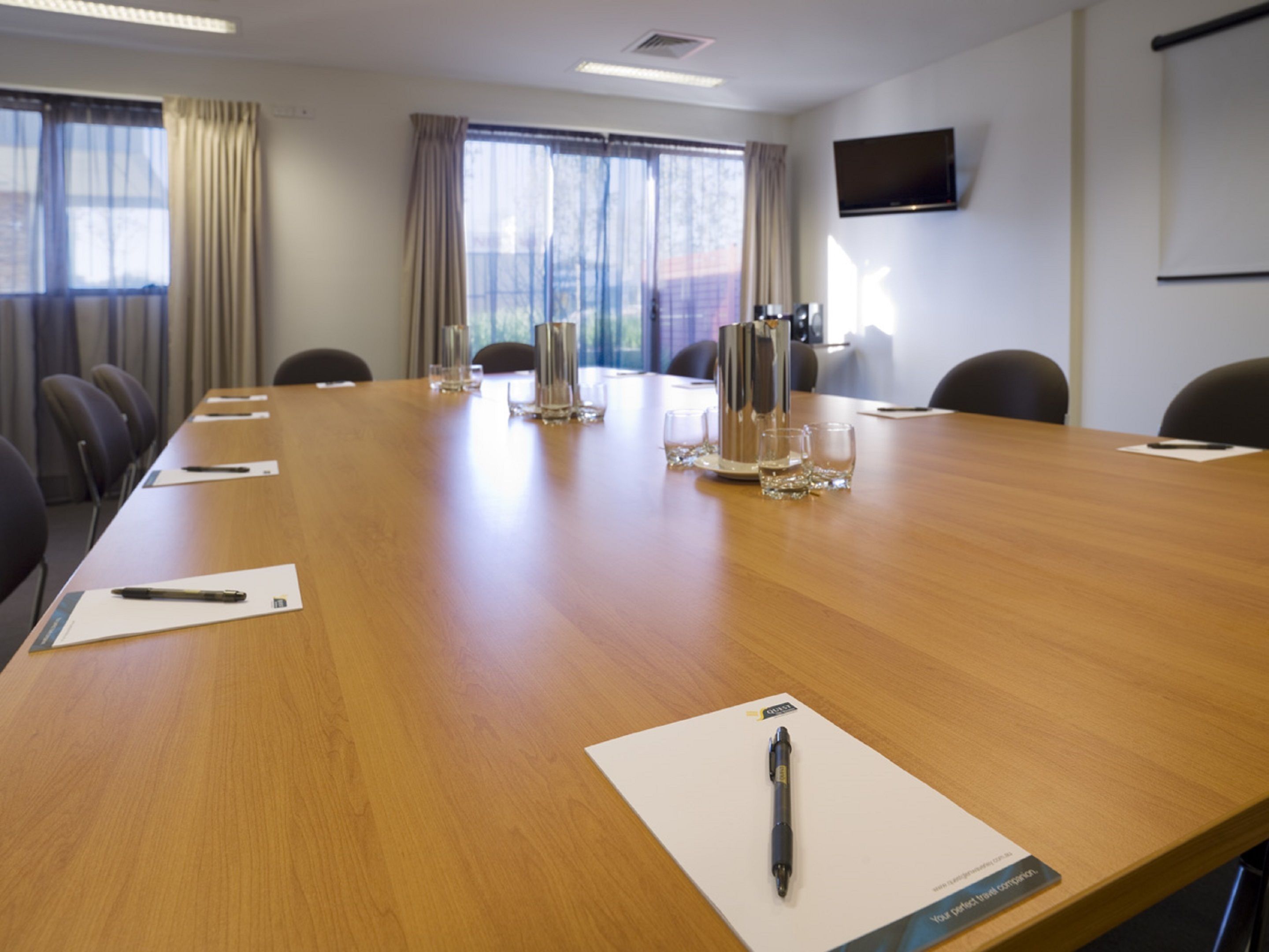 Una sala de conferencias con una larga mesa de madera, sillas, una pantalla de proyector y un televisor en la pared.