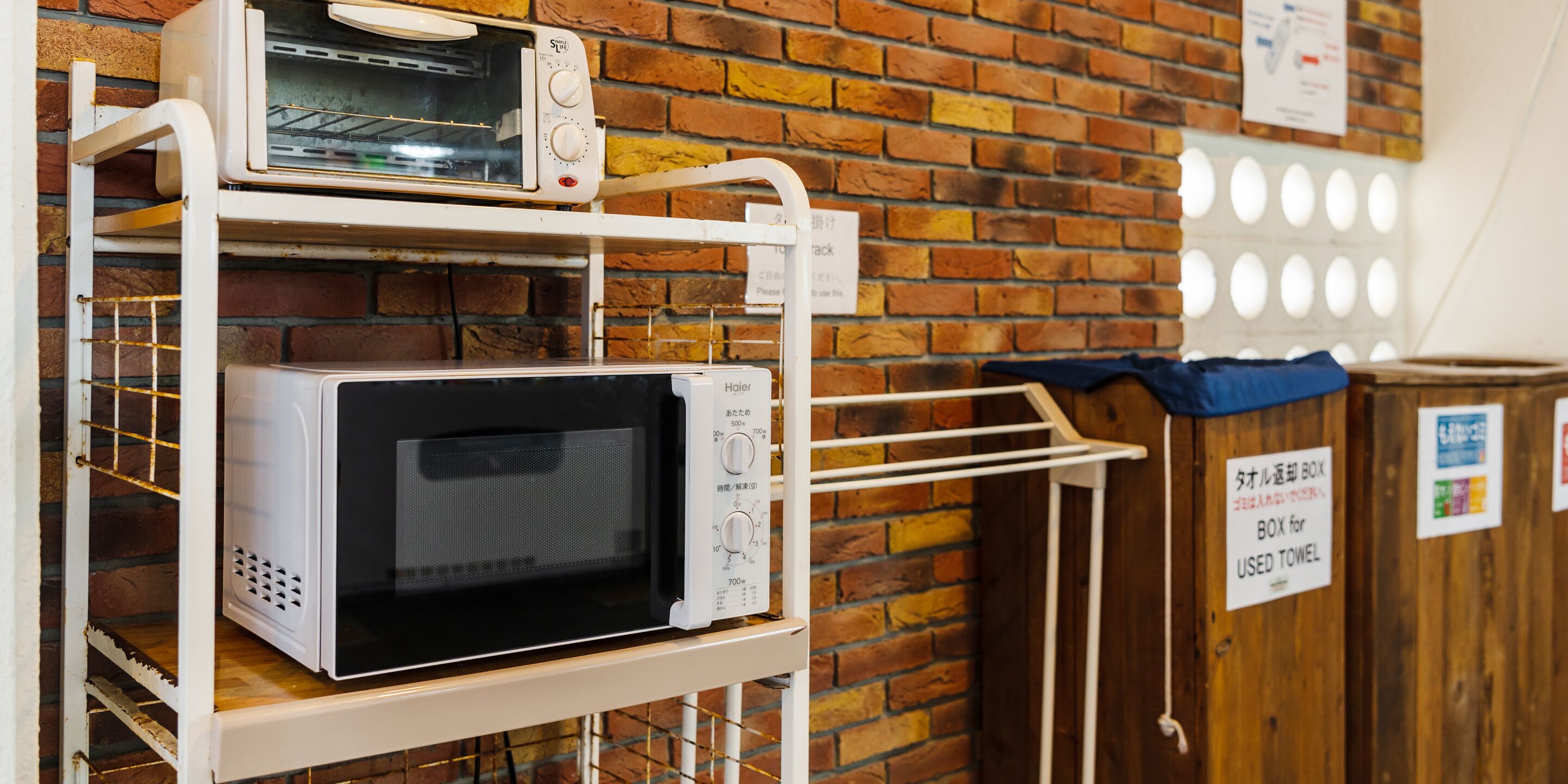 A kitchen area with a brick wall and a microwave oven on a white stand. There is a sign in Japanese on the wall and a blue trash bin.
