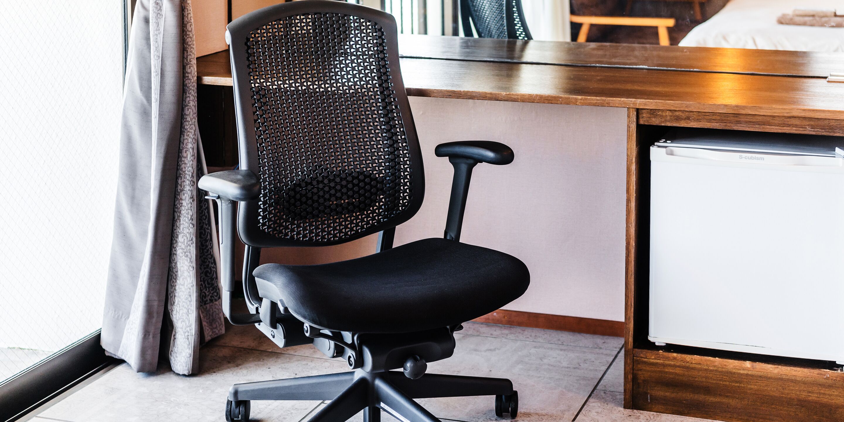 A black office chair with a mesh backrest and armrests, positioned next to a wooden desk and a white refrigerator.