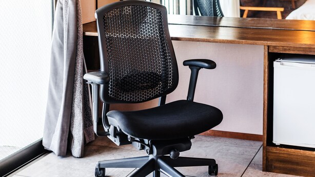 A black office chair with a mesh backrest and armrests, positioned next to a wooden desk and a white refrigerator.