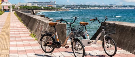 Two bicycles parked on a paved walkway by the sea, with a cityscape in the background.