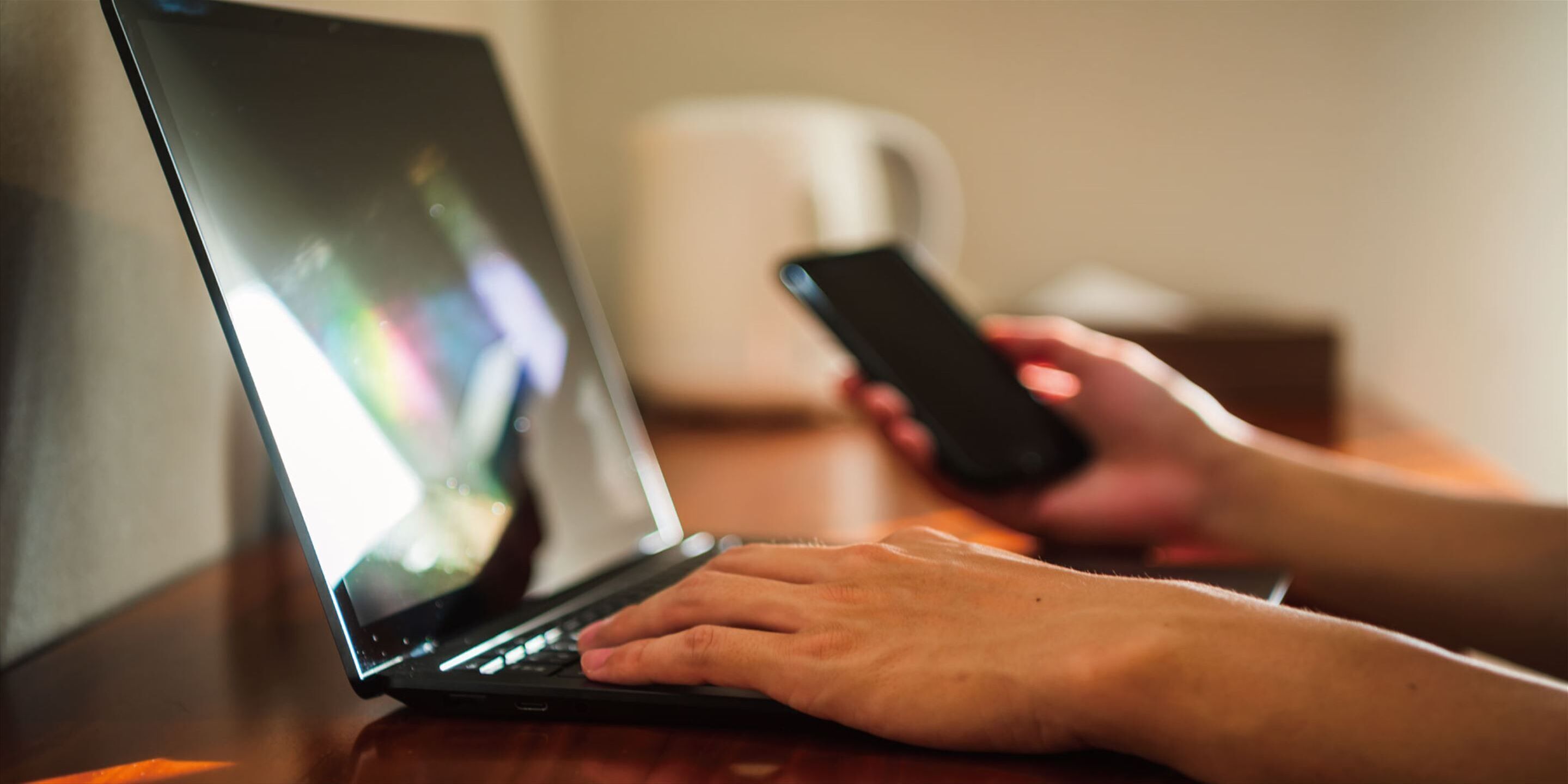 A person using a laptop on a wooden table with a smartphone in hand.