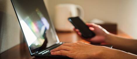 A person using a laptop on a wooden table with a smartphone in hand.