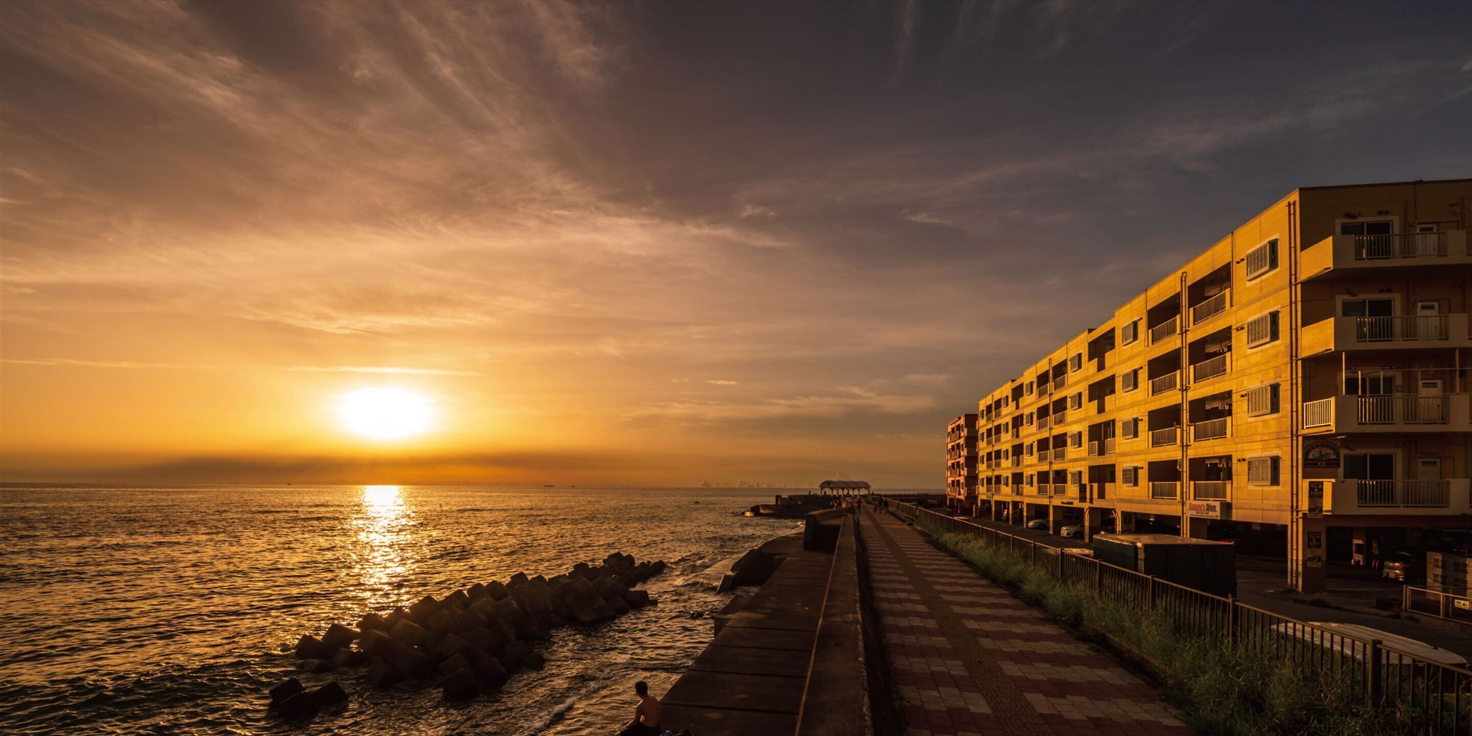 A coastal walkway with a sunset, a multi-story building, and a distant view of the sea.