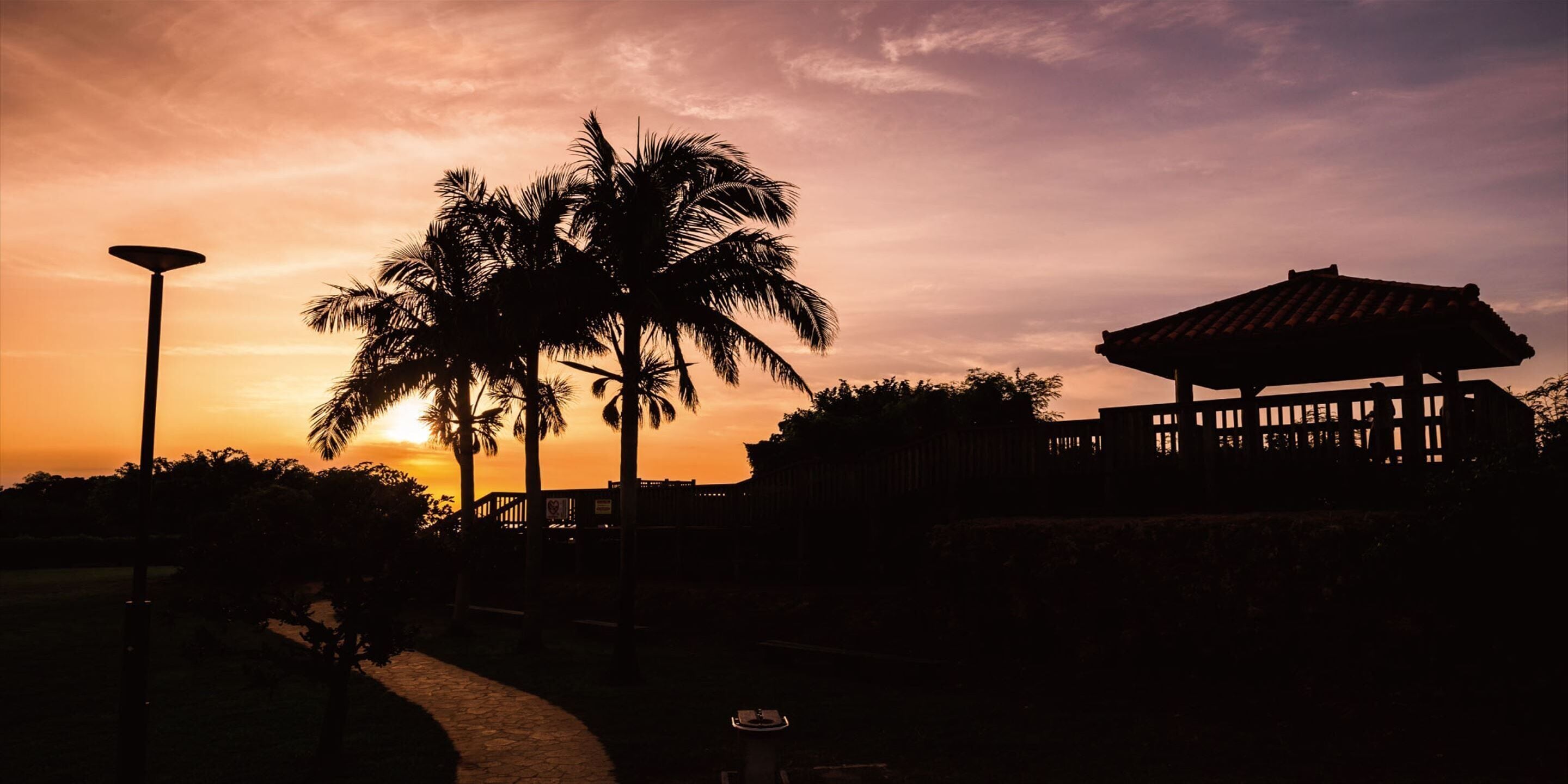 A sunset scene with palm trees, a street lamp, and a building with a tiled roof.