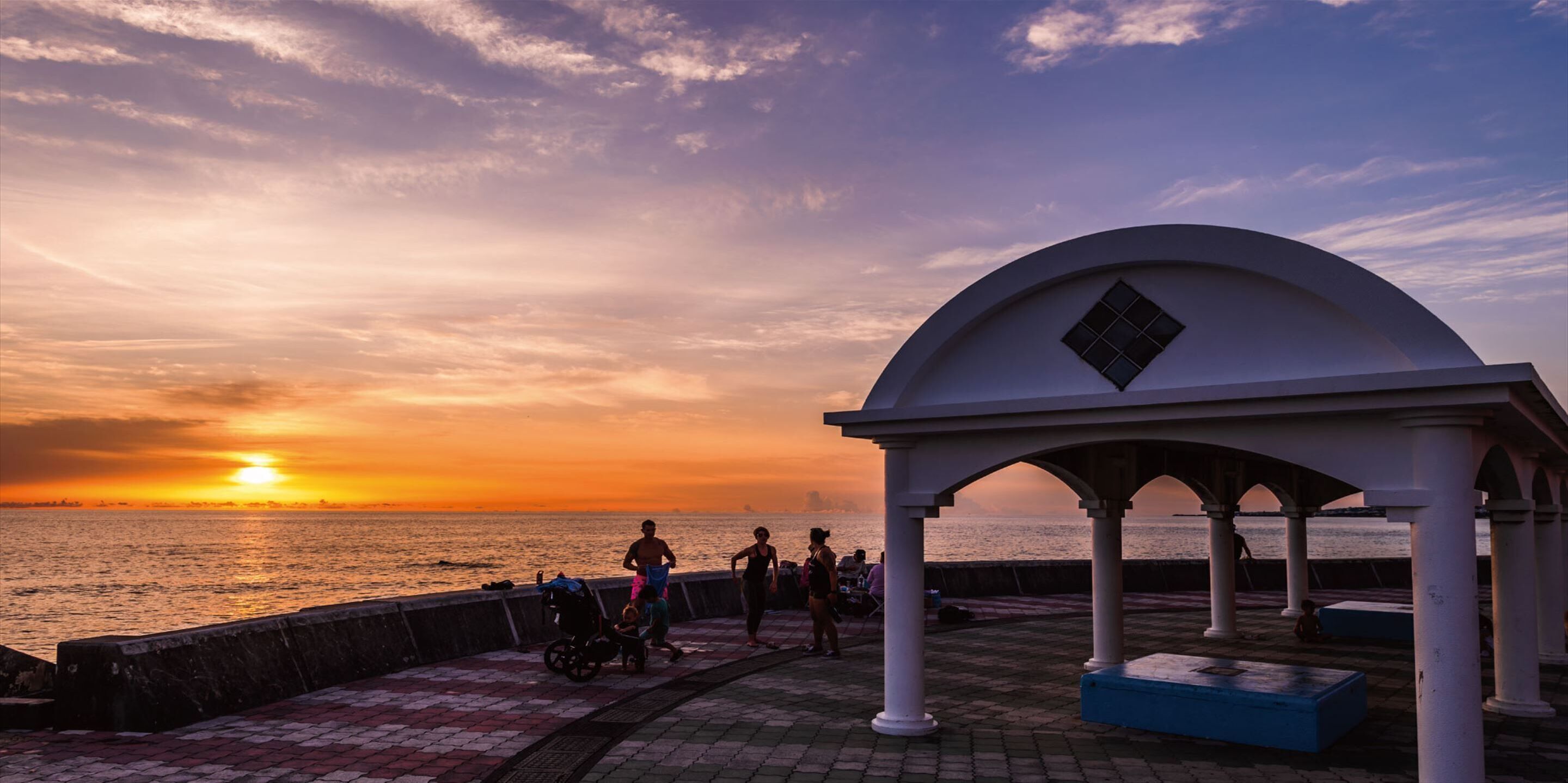 A gazebo with a dome roof overlooking a waterfront during sunset.