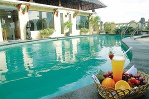 A swimming pool with a drink and fruit basket in the foreground, surrounded by a building with potted plants and a statue.