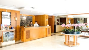 A hotel reception area with a wooden reception desk, a large mirror, and a potted orchid plant.