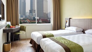 A hotel room with two beds, a desk with a chair, a window with a city view, and a trash can.
