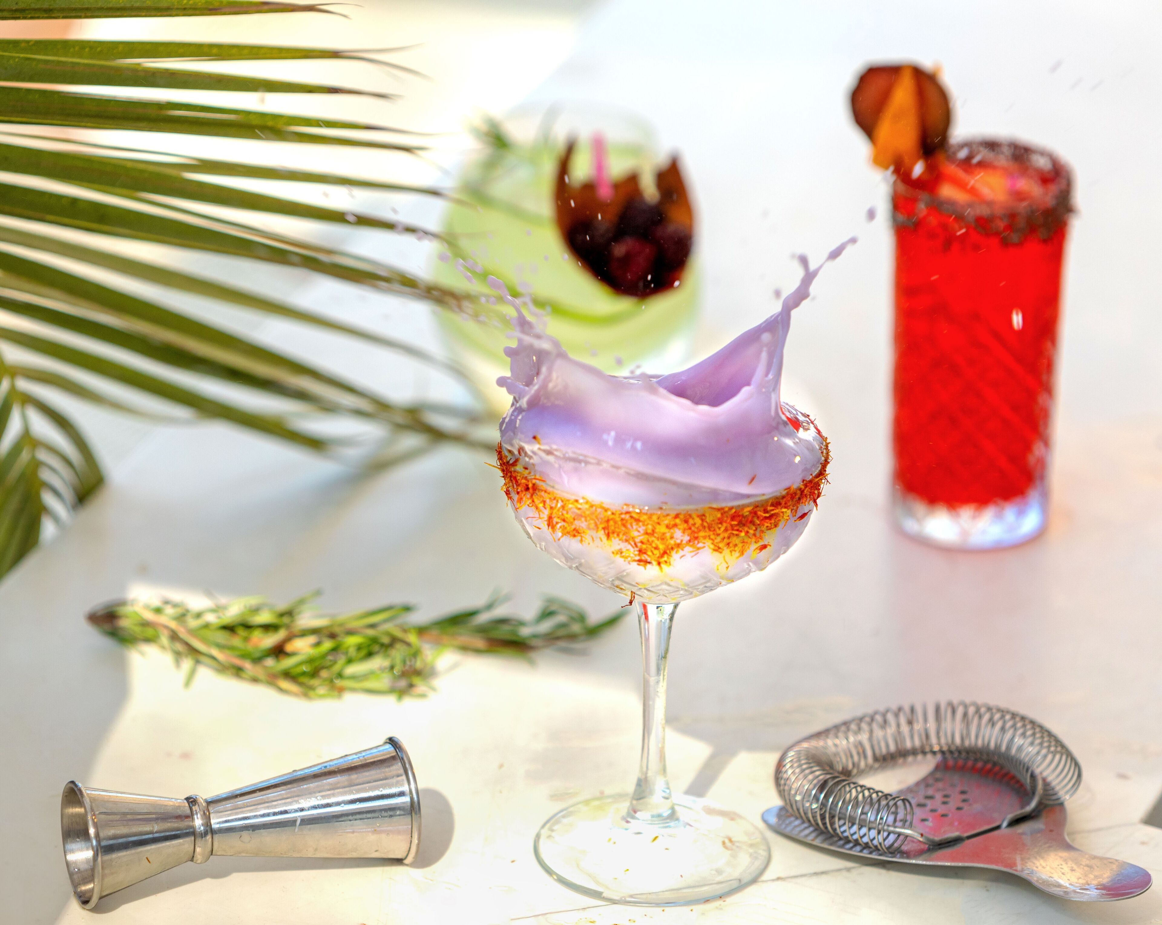 A cocktail glass with a purple liquid splash, a red drink in the background, and a metal strainer on a white surface.