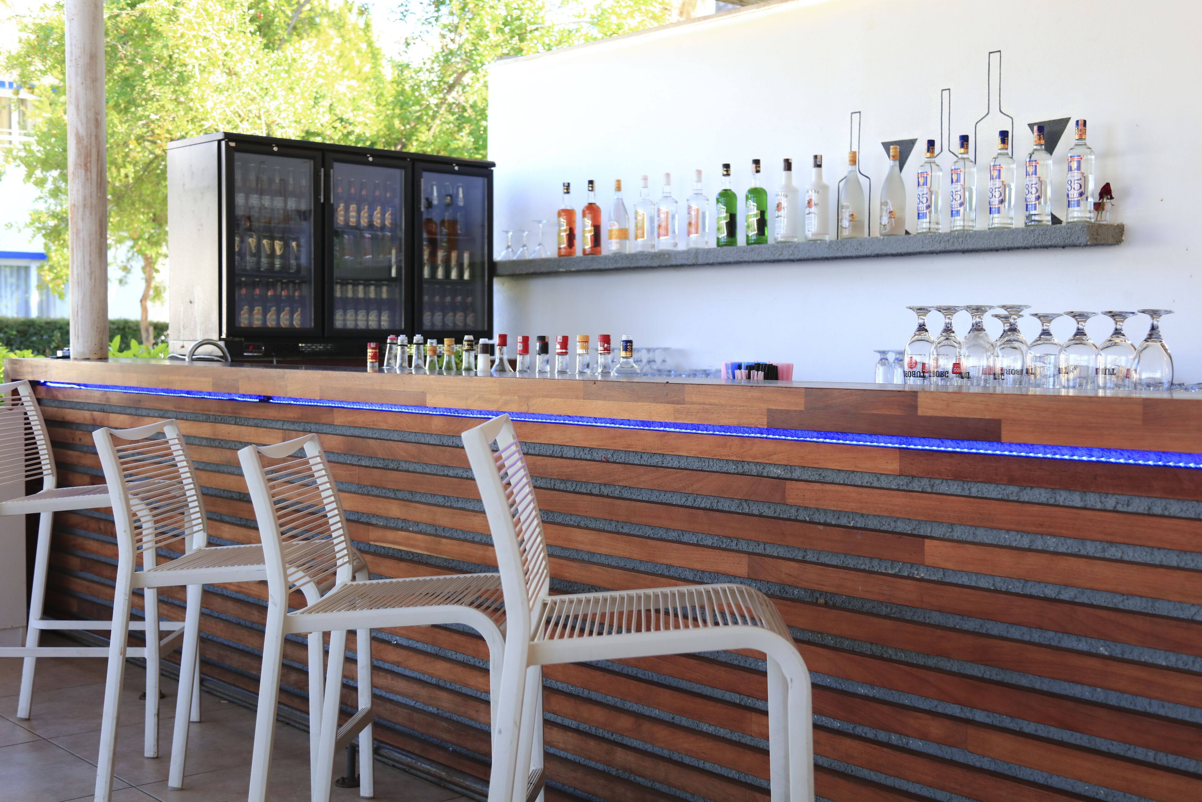 A bar area with a wooden counter, white chairs, and a variety of liquor bottles and glasses.